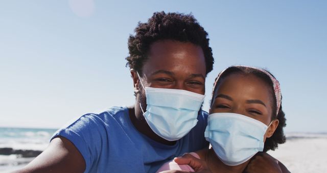 A couple wearing protective masks is happily enjoying a sunny day at the beach. This image portrays positive moments during challenging times, highlighting safety and well-being. Ideal for topics like pandemic lifestyle, safe travel, family moments, and outdoor activities during COVID-19.