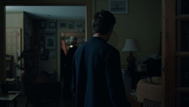 Backview man standing in dimly lit home hallway looking toward gallery wall and lamp