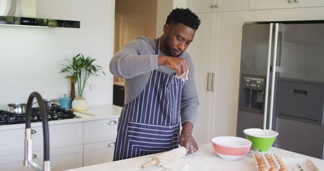 Man in stripe apron baking in modern kitchen, rolling dough and focusing on task. Ideal for cooking blogs, culinary classes, kitchen appliance advertisements, home services.