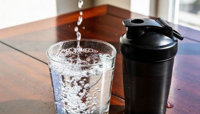 Pouring sparkling water into clear glass beside black protein shaker on wooden table