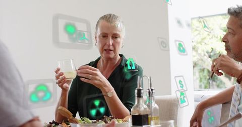 Mature woman leading lively dinner conversation while holding glass at sunlit dining table