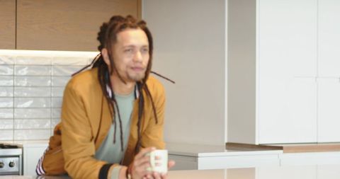 Man with dreadlocks leaning on modern kitchen island holding coffee mug in casual jacket