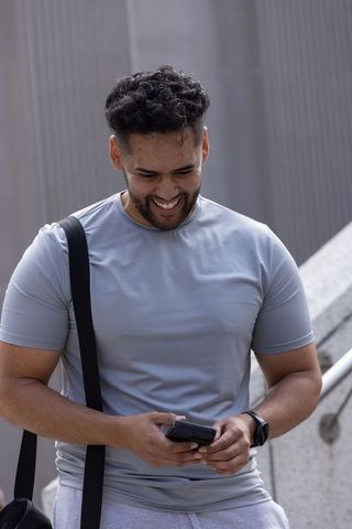 Smiling african american man checking smartphone on urban steps wearing fitted gray shirt