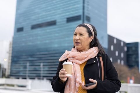Middle-aged woman holding coffee and smartphone outside modern glass office building