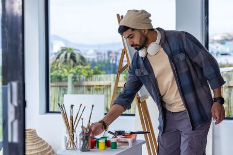 Asian artist arranging paint jars in bright coastal studio wearing beanie and headphones