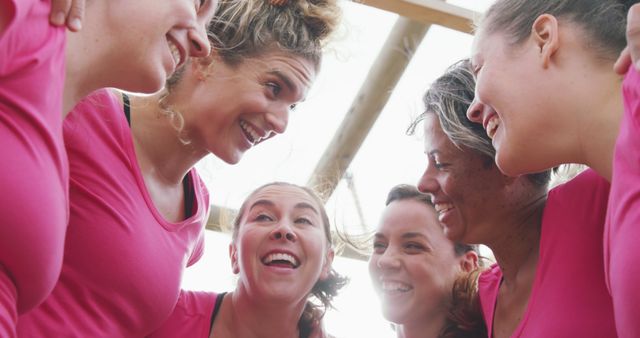 Group of women smiling and bonding outdoors. They are wearing pink shirts and hugging in a friendly circle. This can be used for themes of friendship, teamwork, women's health events, sports activities, and communal support.