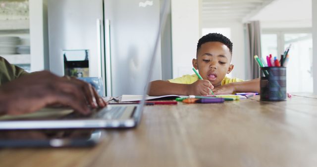 Young boy focused on studying at home with colorful stationery, while a parent works nearby on a laptop. This could be used in articles about balancing work and family life, remote learning, educational resources, or parenting tips. Ideal for promoting home study tools, family dynamics, or telecommuting resources.