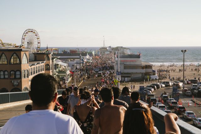 People are walking along a bustling beachfront promenade with an amusement park featuring a ferris wheel in the background. Ocean waves meet a sunny shoreline, creating lively summer atmosphere. Use for vacation promotion, travel guides, recreation ads, summer events marketing.