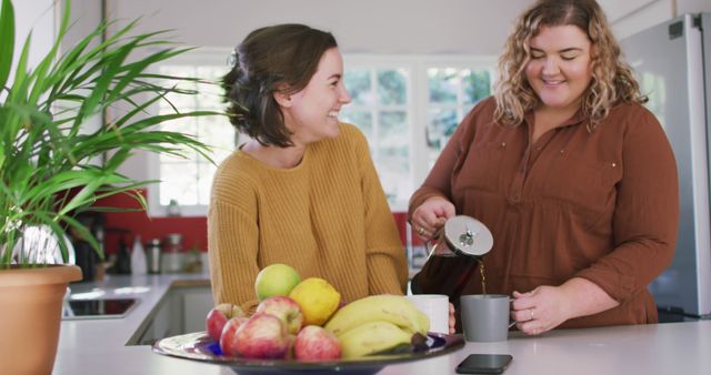 Two women enjoying coffee and laughing together in a bright kitchen. They are showing a strong bond and friendship in a casual home setting with a bowl of fresh fruits on the table. Useful for illustrating friendship, home lifestyle, relaxation, and casual moments with loved ones.