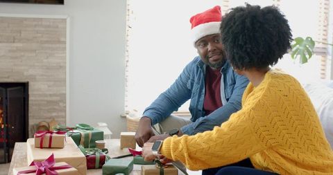 African american couple wrapping christmas gifts and chatting by fireplace in sunlit room