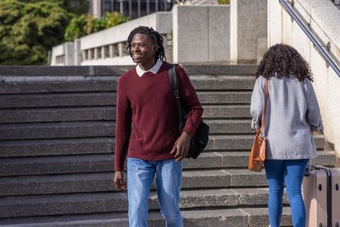 Commuter walking down urban steps smiling with backpack while companion with suitcase