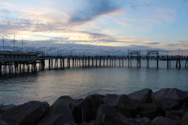 Scenic view of wooden pier stretching into calm sea during sunset, with large rocks in the foreground. Brilliant evening sky with soft, colorful clouds adds tranquility to the scene. Ideal for travel promotions, nature retreats, or relaxing landscape displays.