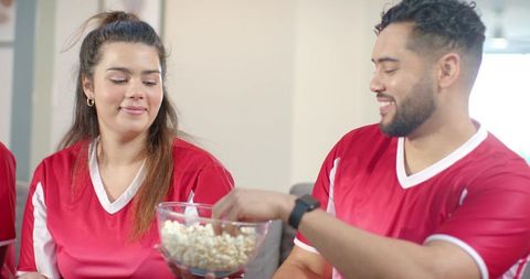 Friends sharing popcorn while watching soccer game in matching red jerseys at home
