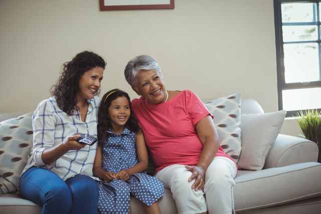 Three generations of women, including a grandmother, mother, and daughter, are sitting on a couch in a living room, smiling and watching television together. This image can be used to depict family bonding, leisure time, and multigenerational relationships. Ideal for advertisements, family-oriented content, and lifestyle blogs.