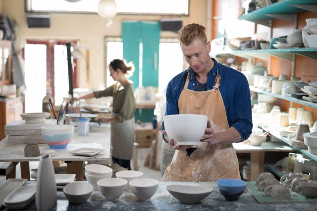 Male potter inspecting a ceramic bowl in a pottery workshop. Shelves filled with various ceramic pieces and pottery tools are visible in the background. Ideal for use in articles about pottery, craftsmanship, small businesses, creative arts, and artisan work.
