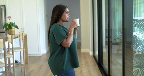 African american woman standing by glass doors holding mug and gazing outside