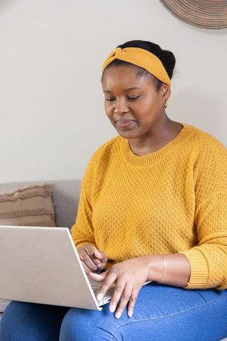 African american woman typing on laptop wearing mustard sweater on cozy sofa