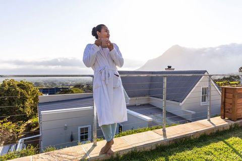 Asian woman in white bathrobe holding mug on rooftop terrace overlooking mountain sunrise