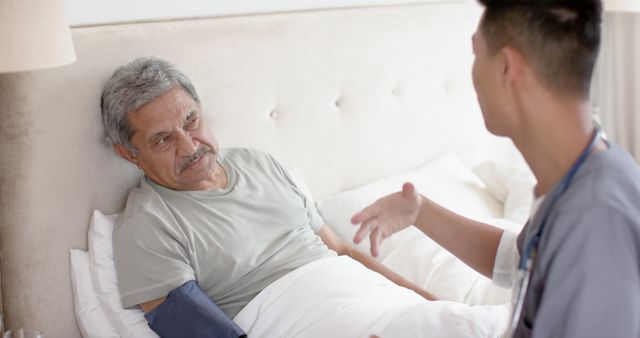 Elderly man lying in bed receiving medical attention from nurse. Nurse discussing care details with patient while monitoring blood pressure. Ideal for use in health care, medical services, home care services, hospital advertisements, and elderly care awareness.