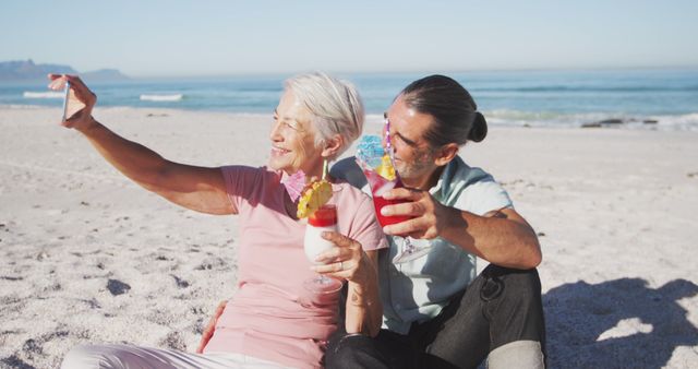 Senior couple sitting on sandy beach, enjoying tropical drinks, and taking selfie. Warm weather, blue ocean, and relaxed atmosphere suggest vacation or holidays. Useful for promoting travel agencies, retirement lifestyle, and holiday activities.