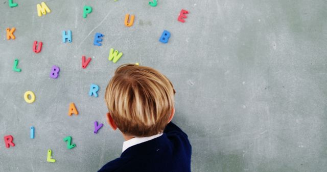 Child wearing school uniform engaging with colorful magnetic letters positioned on a chalkboard. Perfect for educational content, schooling blogs, early childhood development articles, and back-to-school campaigns.