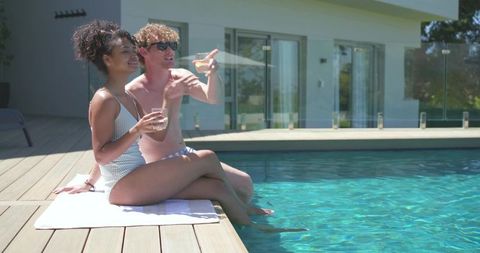 Couple relaxing poolside drinking cocktails dipping feet on sunny modern deck