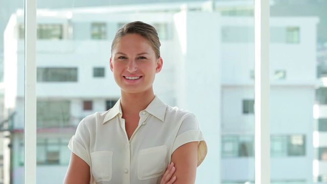 Young businesswoman standing with arms crossed in a bright office, smiling confidently. Ideal for use in presentations, professional blogs, business websites, corporate brochures, or advertisements promoting leadership and professional services.