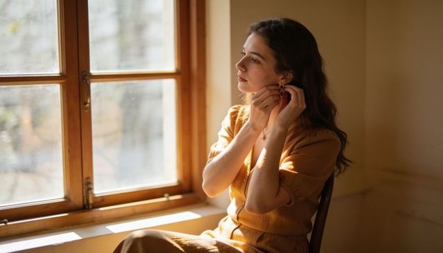 Sunlit woman adjusting gold earring by wooden window in mustard dress