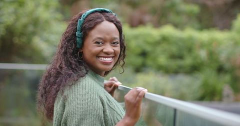 Smiling african american woman leaning on glass balcony railing wearing green sweater