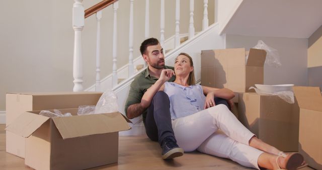 Young couple sitting on floor among moving boxes in new house, appearing relaxed. They are smiling and looking content. Ideal for illustrating themes of moving house, home improvement, relationships, new beginnings, and lifestyle changes.
