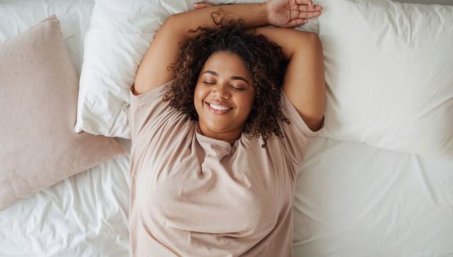 Sunlit morning bliss woman stretching on bed smiling with eyes closed wearing beige tee