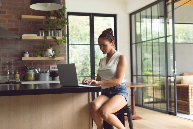 Woman sitting on a stool in a modern kitchen, using a laptop on the counter. Relaxed and casual atmosphere highlights work-from-home lifestyle, suitable for articles and advertisements related to remote work, home office setup, modern living, and technology use.