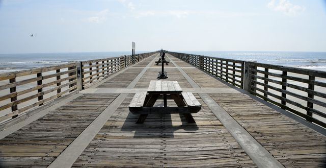 Empty wooden boardwalk stretching into distance over ocean, featuring picnic tables aligned symmetrically. Calm and serene seascape ideal for travel blogs, vacation advertisements, and beach-themed projects.