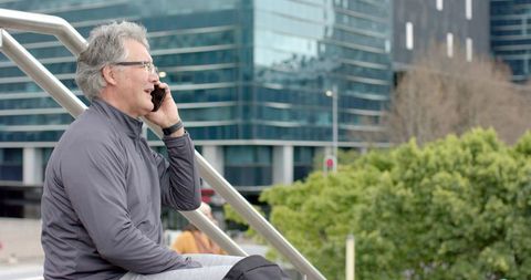 Mature man sitting on modern plaza steps talking on mobile phone wearing knee brace, cityscape