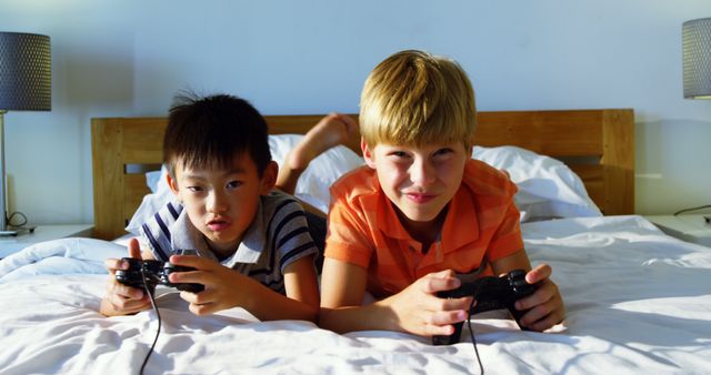 Two boys of different ethnicities are having fun playing video games on a bed, using controllers and smiling. Perfect for concepts related to childhood, gaming, bonding, friendship, and leisure activities in home environment.