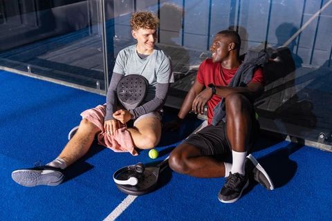 Young diverse friends resting on paddle tennis court sharing laugh after match
