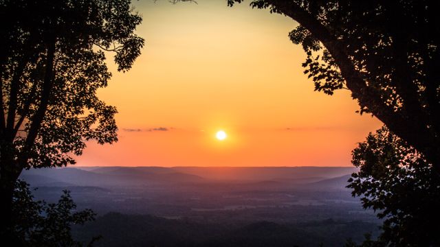 Sun setting over a mountainous landscape with tree silhouettes framing the view. Great for backgrounds, nature-themed projects, travel brochures, relaxation imagery, and inspirational content.