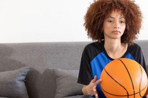 African american woman holding basketball on gray sofa, contemplative athlete at home