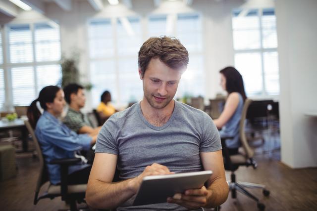Young professional using tablet in modern office with colleagues in background. Ideal for illustrating modern work environments, technology in business, coworking spaces, and teamwork. Suitable for business, technology, and productivity themes.