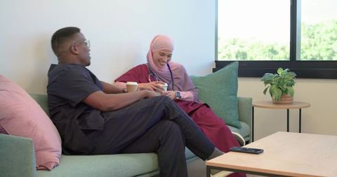 African american clinician and hijab-wearing coworker chatting on lounge sofa with coffee