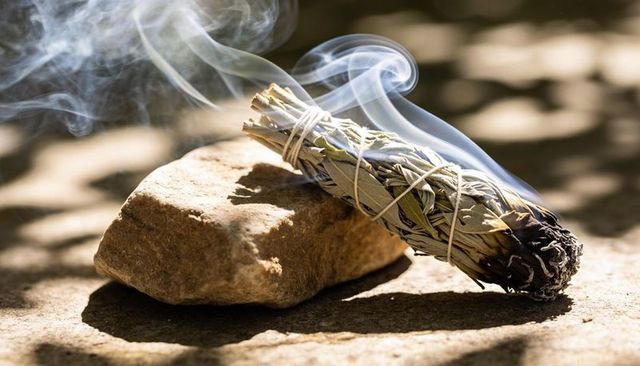 Smoldering white sage smudge stick releasing wisps of smoke on sunlit natural stone