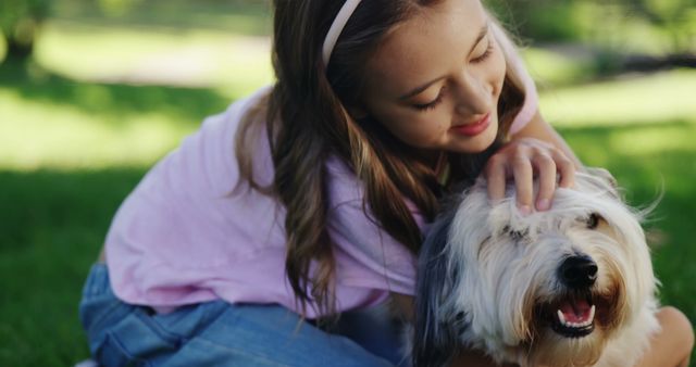 A young girl is affectionately petting a happy dog while sitting on the grass in a park, with copy space. Her gentle interaction with the pet showcases a bond between humans and animals.