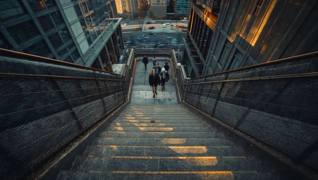 Commuters climbing sunlit staircase between towering glass skyscrapers at golden hour