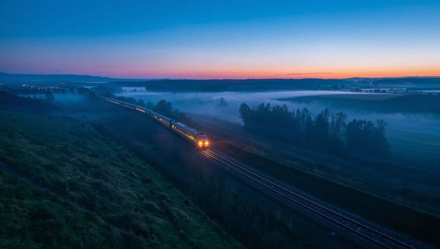 Diesel passenger train cutting through morning fog in rural valley at sunrise golden light