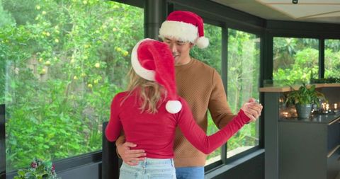 Couple dancing and holding hands in sunroom wearing santa hats, romantic holiday moment