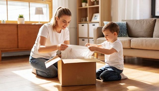 Mother and son unpacking cardboard box in sunlit living room, sharing cozy family moment
