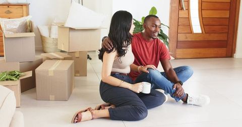 Couple sitting on floor unpacking moving boxes in bright modern home, sharing coffee