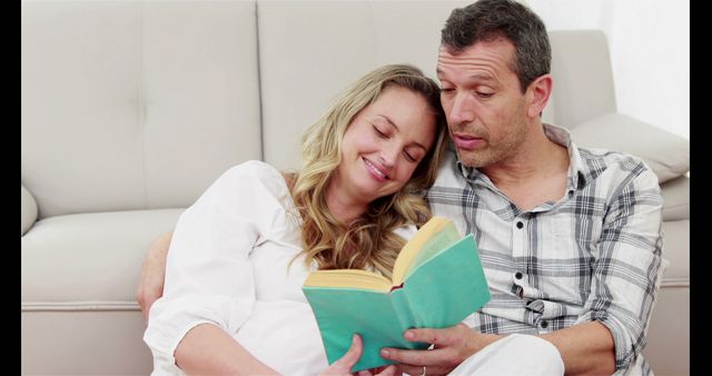 Husband reading book with his pregnant wife in living room