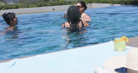 Three women socializing in pool, relaxing with cocktail and smartphone on towel