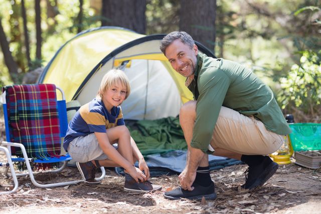 Father and son tying shoelaces in front of a tent at a campsite. Ideal for promoting family bonding, outdoor activities, camping gear, and nature vacations. Perfect for use in advertisements, brochures, and websites related to family travel, outdoor adventures, and parenting.
